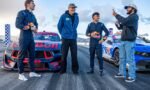 Four men in racing suits stand beside two Red Bull race cars, smiling and chatting on a trackside moment after a run.