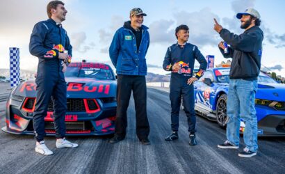 Four men in racing suits stand beside two Red Bull race cars, smiling and chatting on a trackside moment after a run.