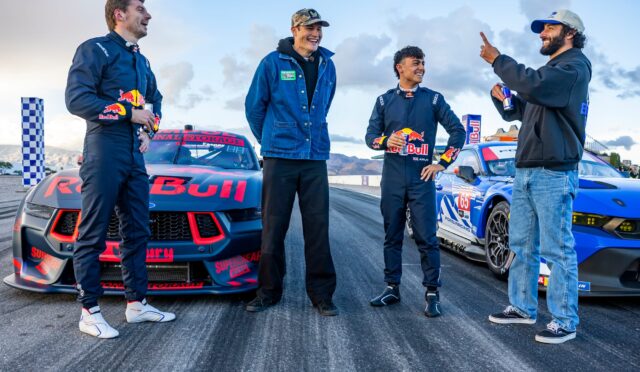 Four men in racing suits stand beside two Red Bull race cars, smiling and chatting on a trackside moment after a run.