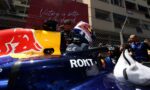 Front view of a Red Bull Racing Formula 1 car in the pit lane, driver with helmet seated while crew members work nearby; chain-link fence and a red banner are in the background.