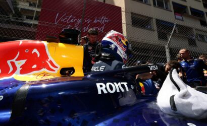 Front view of a Red Bull Racing Formula 1 car in the pit lane, driver with helmet seated while crew members work nearby; chain-link fence and a red banner are in the background.