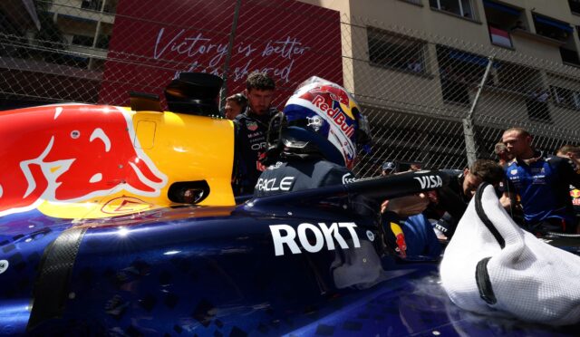 Front view of a Red Bull Racing Formula 1 car in the pit lane, driver with helmet seated while crew members work nearby; chain-link fence and a red banner are in the background.