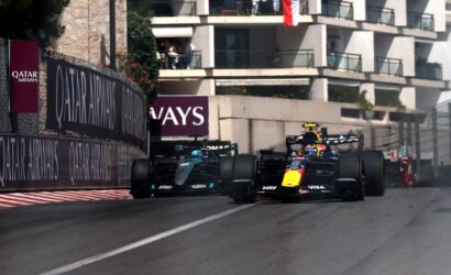 Two Formula 1 cars race side by side on a city street circuit with sponsor barriers and a building backdrop, Qatar Airways banners visible.