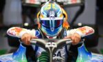 Front view of a race car driver wearing a colorful helmet and blue racing suit, hands gripping the steering wheel in the cockpit.