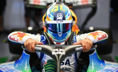 Front view of a race car driver wearing a colorful helmet and blue racing suit, hands gripping the steering wheel in the cockpit.