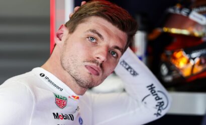 Close-up of a male race car driver in a white suit with Mobil 1 and TAG Heuer logos, tilting his head back against a red wall.