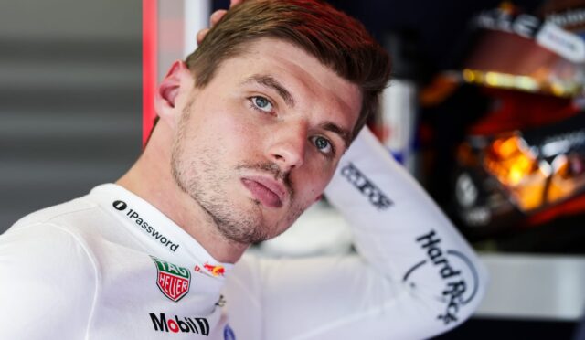 Close-up of a male race car driver in a white suit with Mobil 1 and TAG Heuer logos, tilting his head back against a red wall.