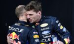 Two race car drivers in blue sponsor suits hug on a dark backdrop, Red Bull logo visible on their outfits.