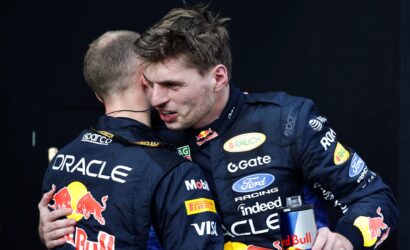 Two race car drivers in blue sponsor suits hug on a dark backdrop, Red Bull logo visible on their outfits.