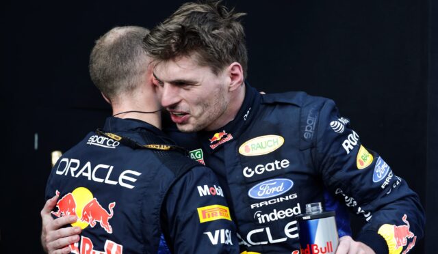 Two race car drivers in blue sponsor suits hug on a dark backdrop, Red Bull logo visible on their outfits.