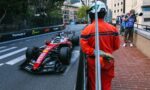Red Formula 1 car on a Monaco street circuit, flanked by barriers as photographers and an official in an orange suit watch from the sidewalk.