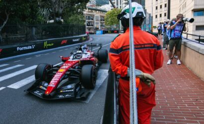 Red Formula 1 car on a Monaco street circuit, flanked by barriers as photographers and an official in an orange suit watch from the sidewalk.
