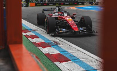 Red Ferrari Formula 1 car on a race track, framed by orange barriers in the foreground as it turns a corner.