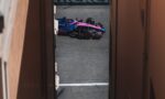 Pink and blue Formula 1 car peeks through a narrow gap between orange walls on a racetrack driving surface.