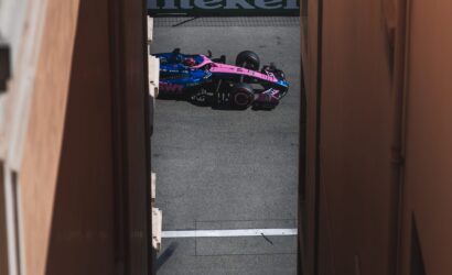 Pink and blue Formula 1 car peeks through a narrow gap between orange walls on a racetrack driving surface.