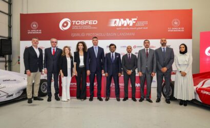Group of professionals in formal attire posed for a photo at a press event with red banners and sponsor logos behind them in a showroom/wrapper area.