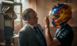 Man in a gray suit helps a race car driver adjust a bright, multicolored helmet.