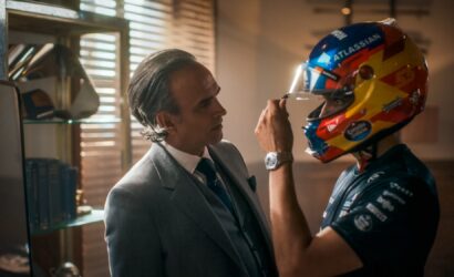 Man in a gray suit helps a race car driver adjust a bright, multicolored helmet.