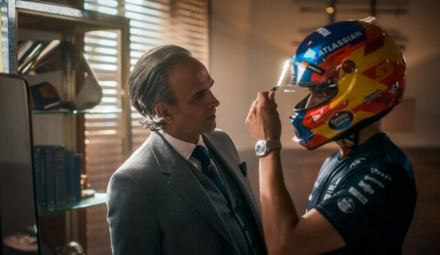 Man in a gray suit helps a race car driver adjust a bright, multicolored helmet.