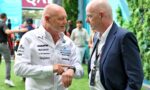 Two bald men conversing at an outdoor event; one in a white sponsor shirt, the other in a navy suit with lanyards nearby.