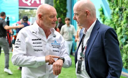 Two bald men conversing at an outdoor event; one in a white sponsor shirt, the other in a navy suit with lanyards nearby.