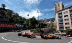 Formula 1 cars speed around a tight Monaco street circuit with spectators on balconies and a tall pink apartment building on the right.