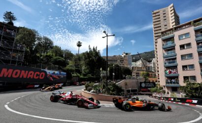 Formula 1 cars speed around a tight Monaco street circuit with spectators on balconies and a tall pink apartment building on the right.