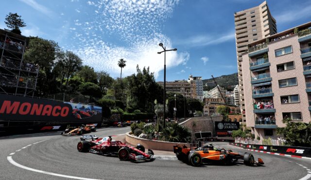 Formula 1 cars speed around a tight Monaco street circuit with spectators on balconies and a tall pink apartment building on the right.