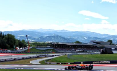 Race car with Oracle Red Bull livery speeding around a track with mountains in the background, under a blue sky.