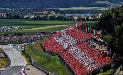 Massive stadium crowd in red and white fills the grandstands at a racing circuit, with a curving track on the left and green fields beyond.