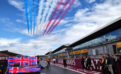 Jet aerobatic team flies overhead, trails of red, white, and blue smoke above a racetrack grandstand and pit building.",