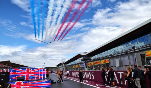 Jet aerobatic team flies overhead, trails of red, white, and blue smoke above a racetrack grandstand and pit building.",