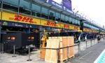 Pit lane at a race track with DHL banners, McLaren branding, and equipment crates visible along the pit wall in front of glass-fronted garages.