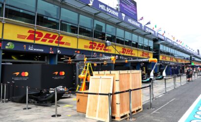 Pit lane at a race track with DHL banners, McLaren branding, and equipment crates visible along the pit wall in front of glass-fronted garages.
