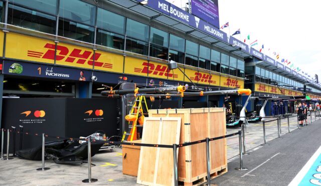 Pit lane at a race track with DHL banners, McLaren branding, and equipment crates visible along the pit wall in front of glass-fronted garages.