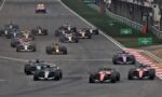 A pack of Formula 1 cars speeds down a race track, close together near the pit wall and cheering spectators in the background.
