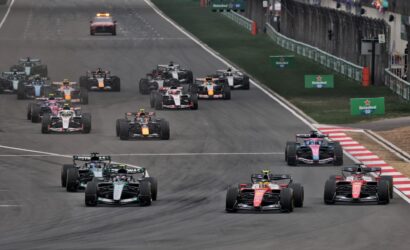 A pack of Formula 1 cars speeds down a race track, close together near the pit wall and cheering spectators in the background.