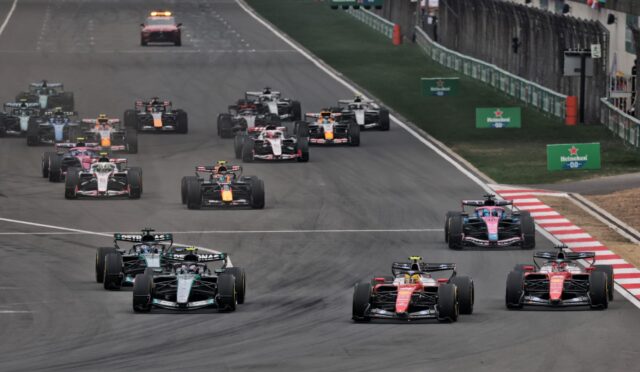 A pack of Formula 1 cars speeds down a race track, close together near the pit wall and cheering spectators in the background.