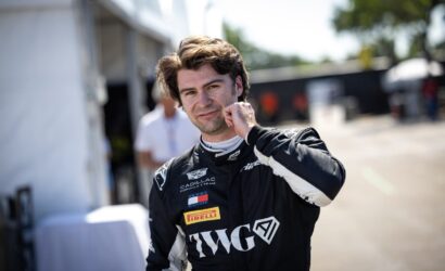 Smiling male race car driver in a black racing suit, touching his cheek in a pit area outside the track.