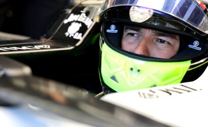 Close-up of a race car driver wearing a neon yellow helmet with a clear visor in a cockpit, helmets branded Schuberth visible.