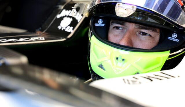 Close-up of a race car driver wearing a neon yellow helmet with a clear visor in a cockpit, helmets branded Schuberth visible.