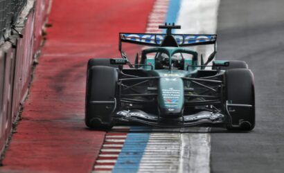 Rear view of a green Formula 1 car racing along a wet track, beside a red barrier and blue-and-white curb.