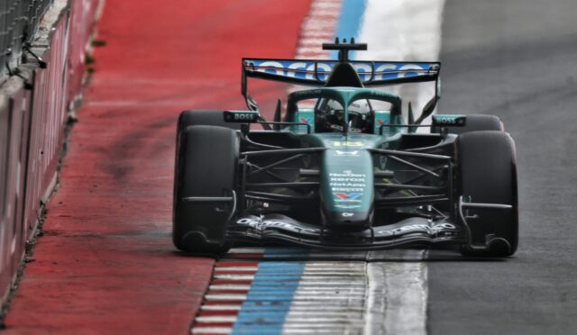 Rear view of a green Formula 1 car racing along a wet track, beside a red barrier and blue-and-white curb.