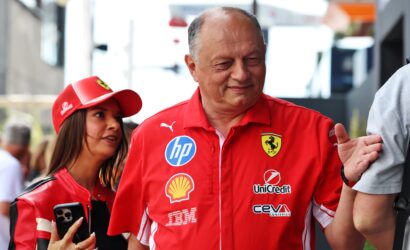 Smiling man in a red Ferrari shirt with sponsor logos, standing outdoors beside a woman in a red cap at a public event.