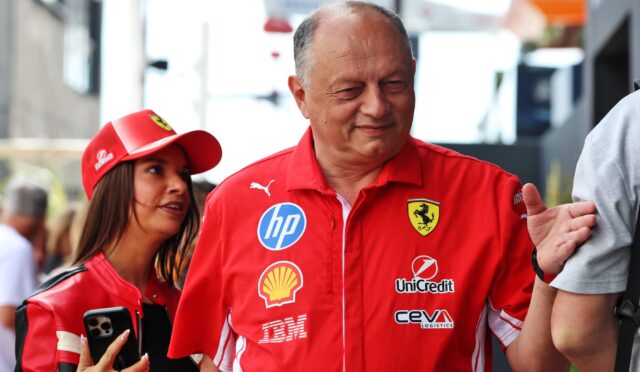 Smiling man in a red Ferrari shirt with sponsor logos, standing outdoors beside a woman in a red cap at a public event.