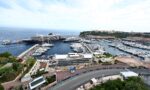 A large cruise ship docked at a busy marina surrounded by many yachts, with a hillside town in the distance.