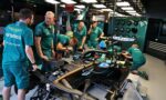 Team of engineers in teal uniforms work on an Aston Martin Formula 1 car inside a well-lit garage.