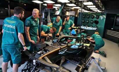 Team of engineers in teal uniforms work on an Aston Martin Formula 1 car inside a well-lit garage.