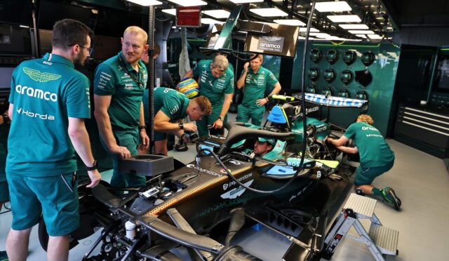 Team of engineers in teal uniforms work on an Aston Martin Formula 1 car inside a well-lit garage.