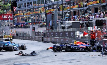 Formula 1 cars stop on a street circuit after a crash, with debris and a damaged front wing on the track and spectators watching from the barriers and grandstands behind safety fencing.